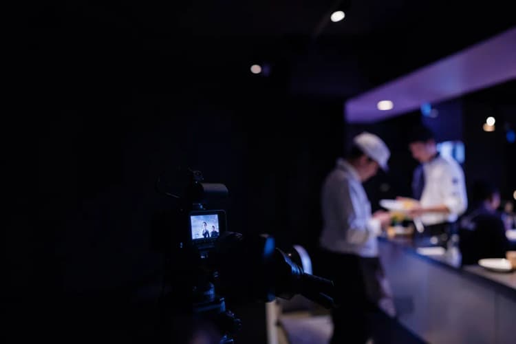 Uma câmera de vídeo profissional filmando um chef preparando um prato em uma cozinha de restaurante iluminada.